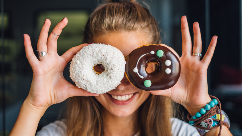 lächelnde junge Frau hält sich zwei bunt dekorierte Donuts vor die Augen