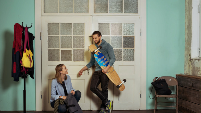 Junges Paar lehnt vor einer Altbauzimmertür, sie packt ihren Rucksack, er hält ein Longboard im Arm