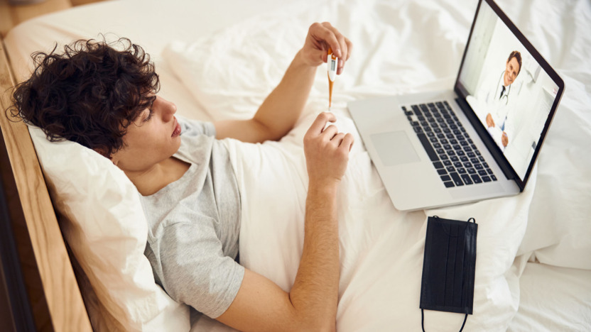 Junge liegt krank im Bett und nimmt eine digitale Sprechstunde über den Laptop war.