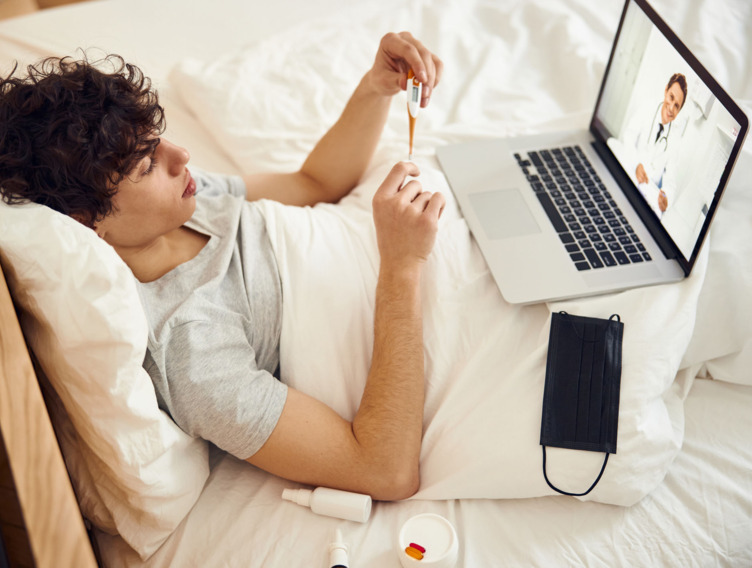 junger Mann liegt im Bett und hat eine Videosprechstunde beim Arzt