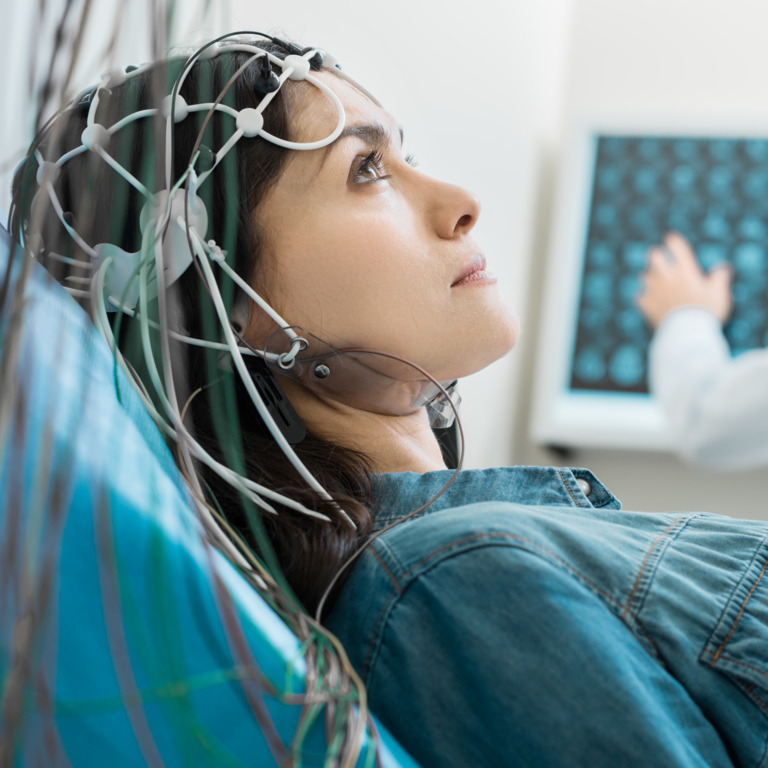 Junge Frau mit Elektroden am Kopf liegt auf einer Behandlungsliege, im Hintergrund sitzt ein Arzt