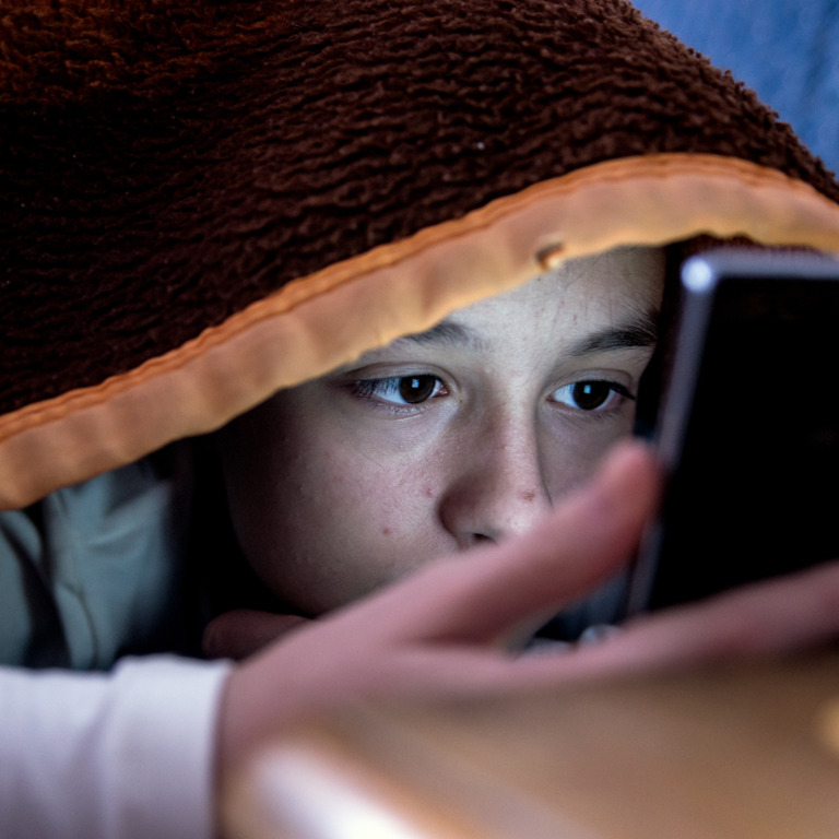 Kind liegt unter der Decke und schaut auf sein Smartphone.