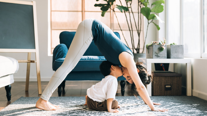 Mutter macht in ihrem Wohnzimmer die Yoga-Übung herabschauender Hund, ihr kleiner Sohn sitzt zwischen ihren Armen und Beinen