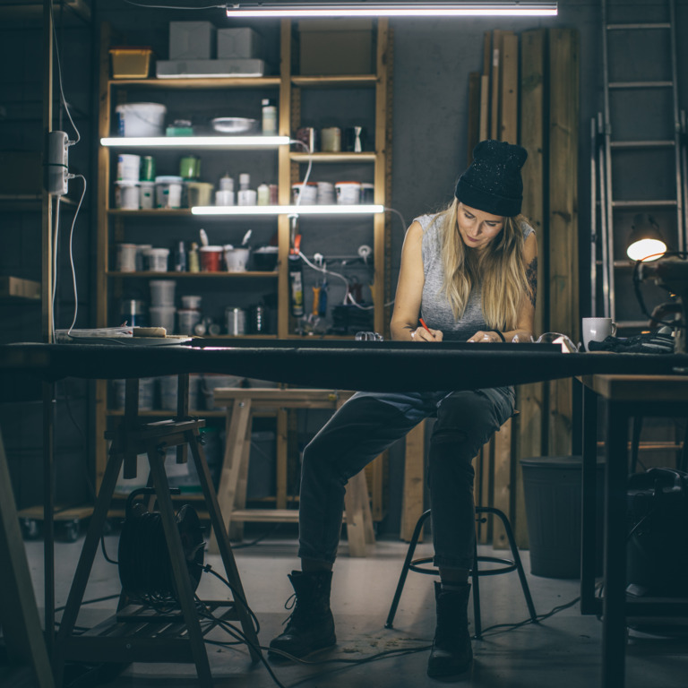 Eine Frau mit Mütze sitzt an einem Tisch in einem Arbeitsraum. Sie hält einen Stift in der Hand und verwendet dieses auf dem Tisch.