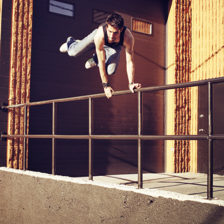 Junger Mann macht Parcouring und springt über ein Geländer auf einem Firmengelände.