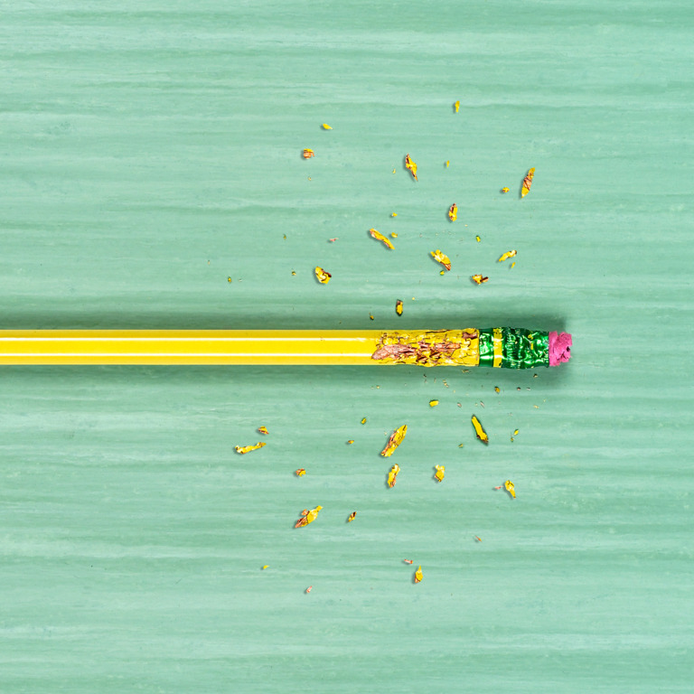 Ein gelber Bleistift mit rotem Radiergummi auf einem grünen Tisch, an dessen oberen Ende Späne vom darauf herumkauen liegen.