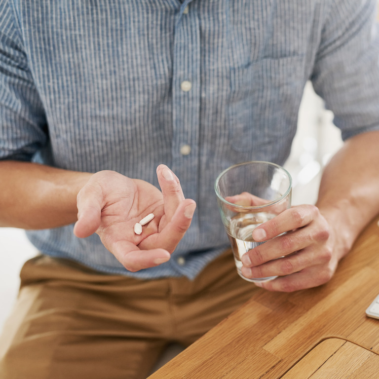 Ein Mann sitzt an seinem Arbeitsplatz und hält ein Glas Wasser in linker Hand und zwei Schmerztabletten in der rechten. 