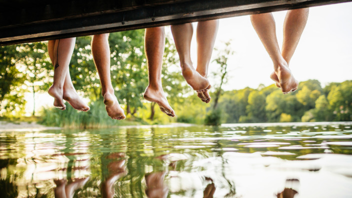 Baumelnde Beine auf einem Steg, die sich im Wasser spiegeln