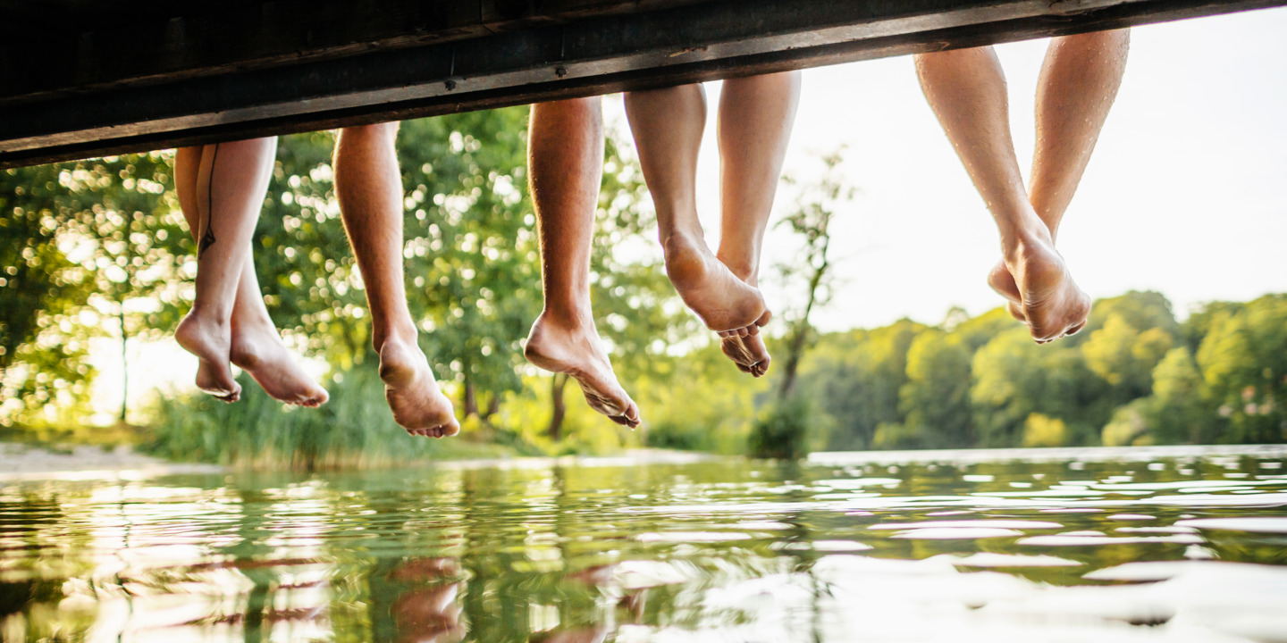 Vier Menschen sitzen an Steg und lassen ihre Beine über dem See baumeln.