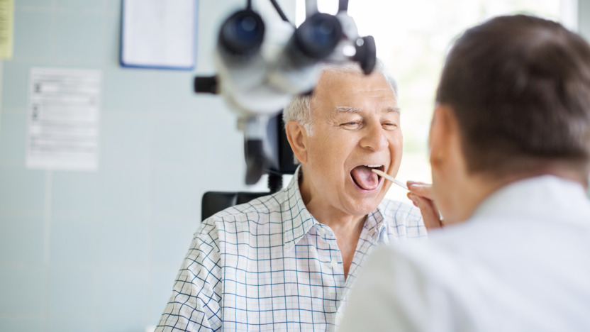 Älterer Herr sitzt mit geöffnetem Mund vor einem Arzt, der mit einem Stäbchen seine Zunge herunterdrückt