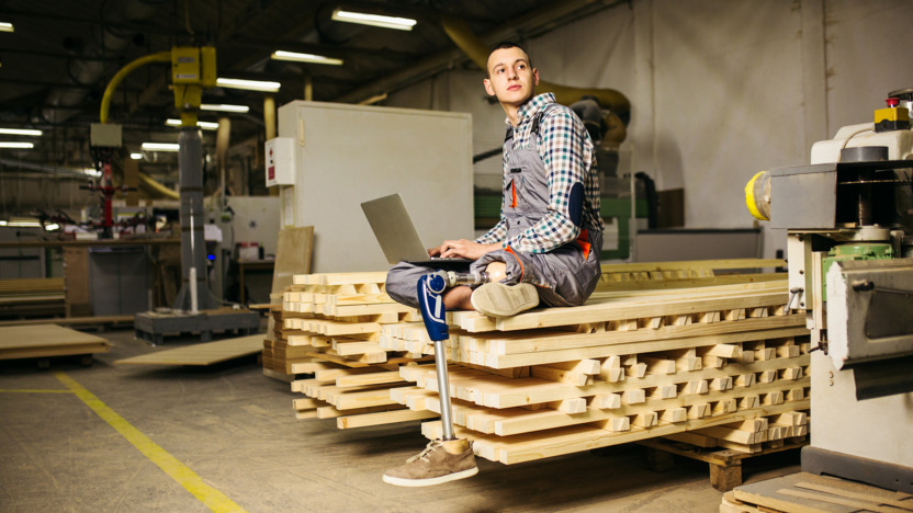 Junger Mann mit Beinprothese sitzt im Schneidersitz auf einem Holzhaufen mit dem Laptop auf dem Schoß