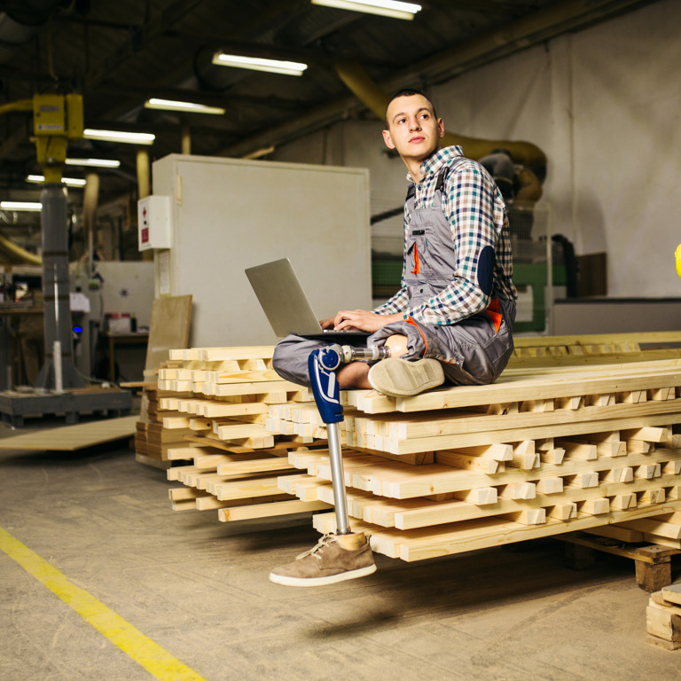 junger Tischler mit einer Beinprothese sitzt mit einem Laptop auf einem Stapel Holzleisten