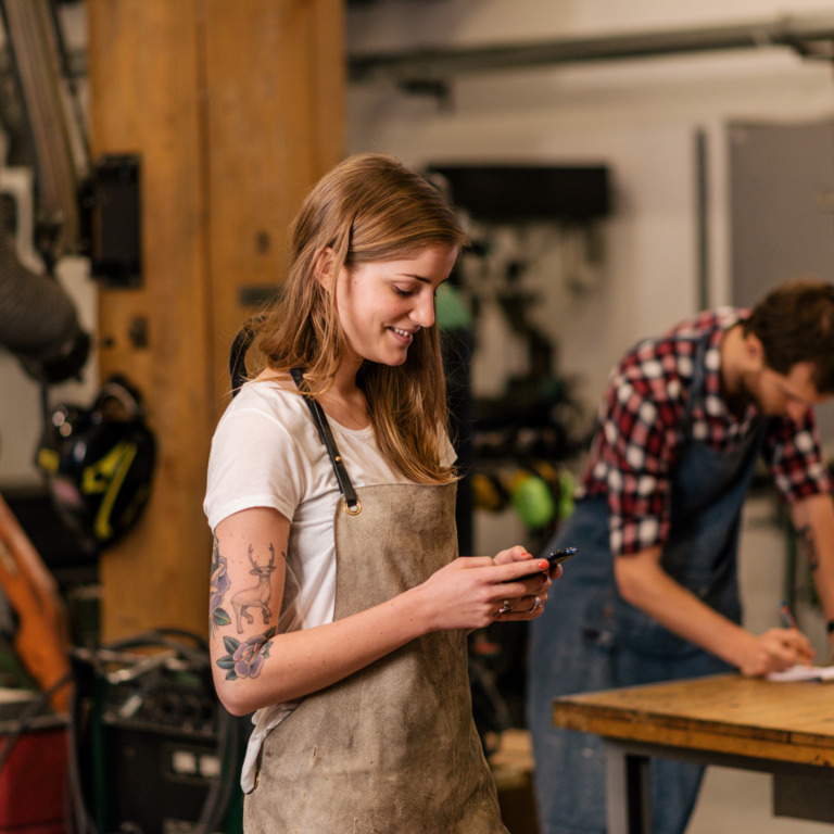 Eine Frau und ein Mann in einer Werkstatt. Die Frau ist am Handy, der Mann arbeitet.