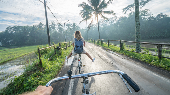 zwei junge Urlauber fahren mit dem Rad zwischen Reisfeldern auf einer kleinen vietnamesischen Landstraße 