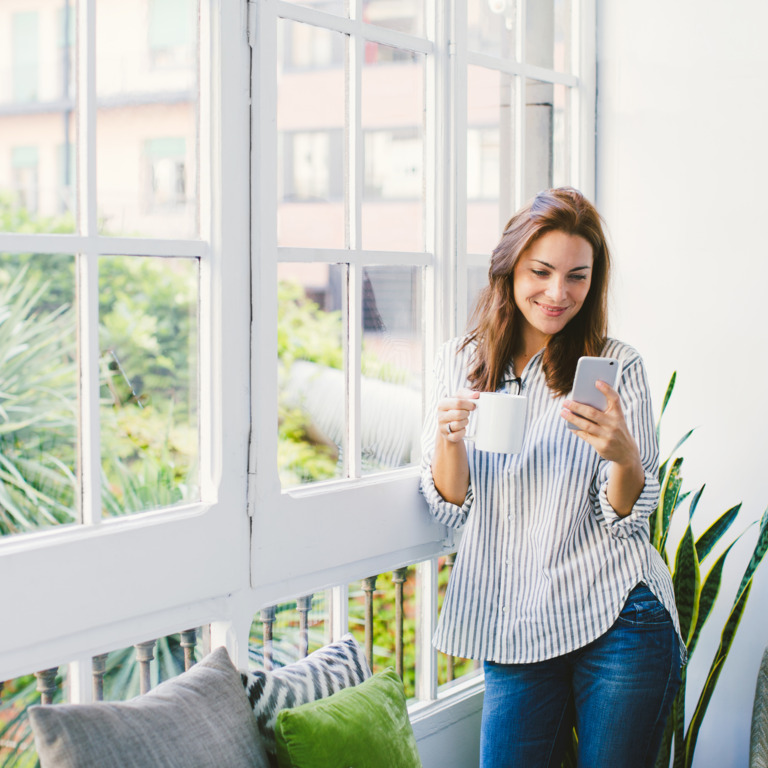 Frau steht mit Smartphone und Tasse am Fenster und blickt lächend auf ihr Handy.