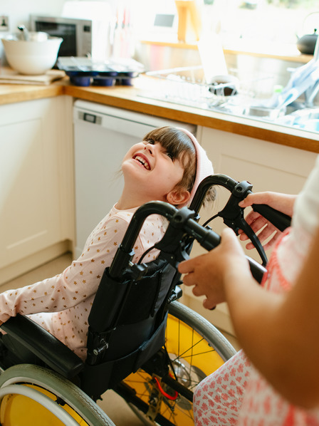 Frau schiebt ein kleines Mädchen im Rollstuhl durch eine Küche