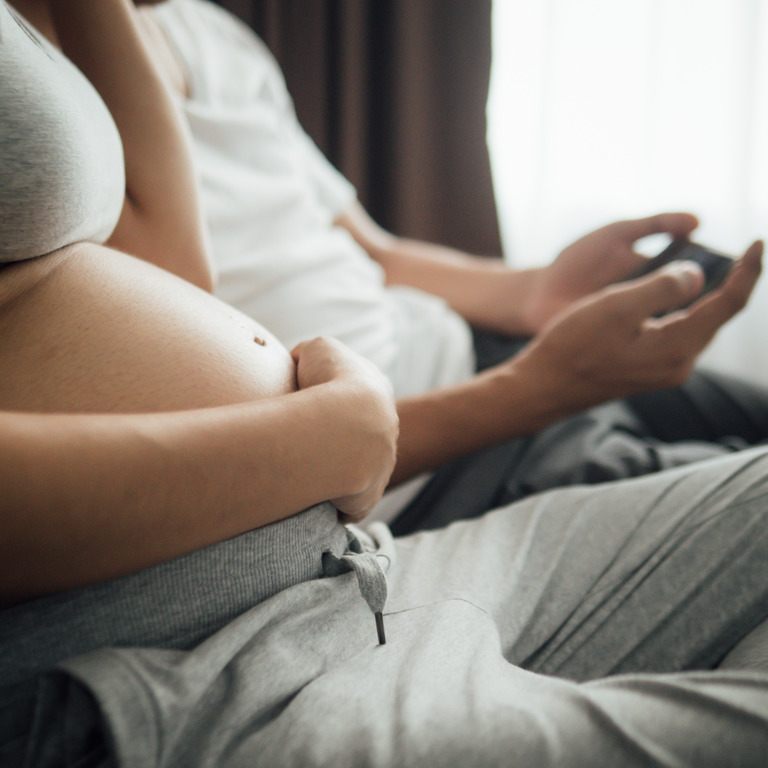 Schwangere und ihr Mann sitzen auf einem Sofa, er checkt sein Smartphone