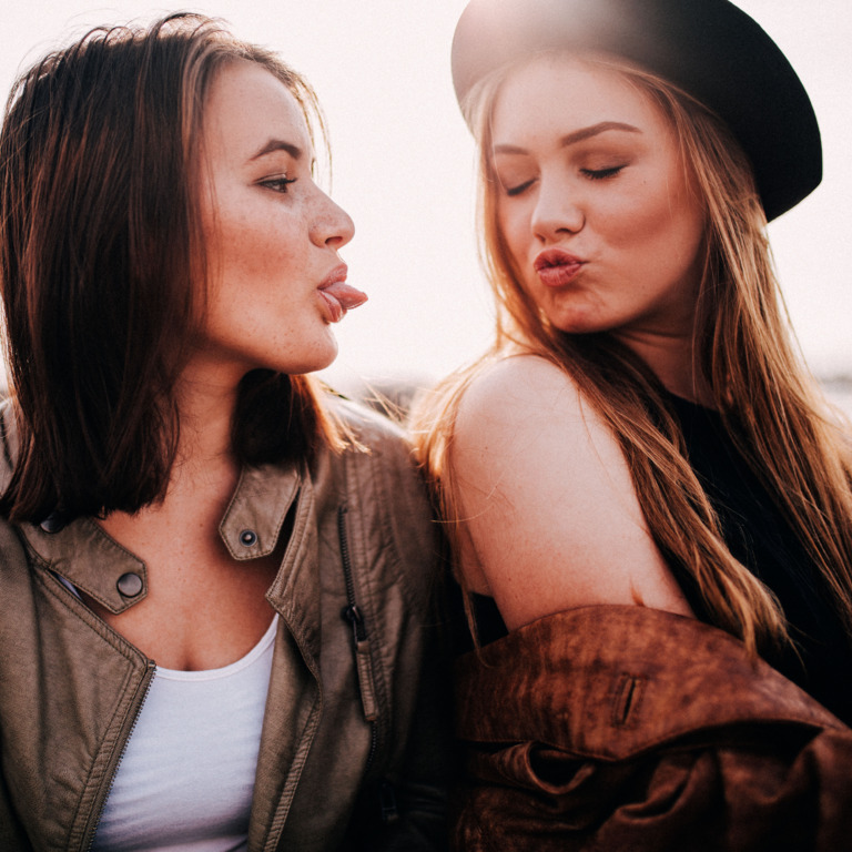 eine Teenagerin macht einen Kussmund in Richtung ihrer Freundin, die Freundin streckt ihr die Zunge raus