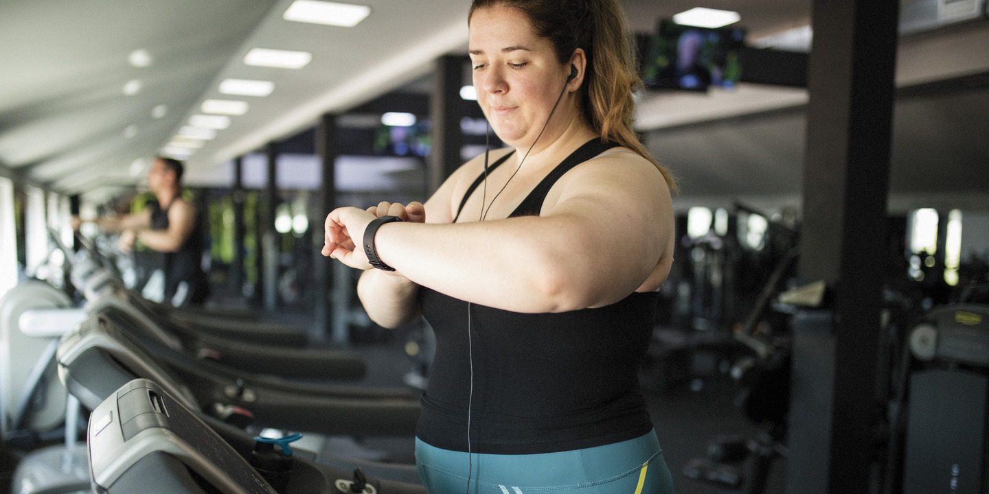 Übergewichtige Frau auf Laufband in Fitnessstudio sieht auf Fitnesstracker an ihrem Handgelenk.