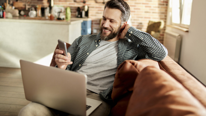 Mann sitzt lächelnd mit Laptop und Handy auf dem Sofa. 