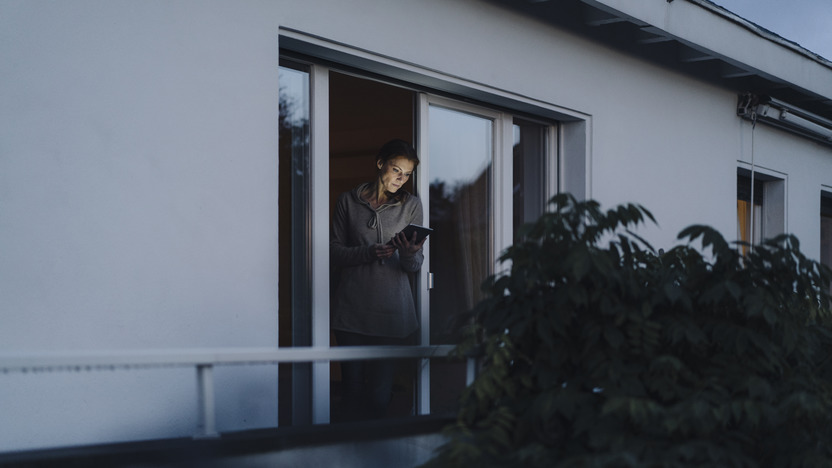 Frau steht in Dämmerung an Balkontür und schaut auf Smartphone
