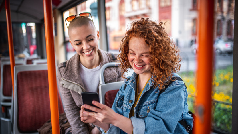 Zwei junge Frauen sitzen im Bus und schauen lachend auf ein Smartphone
