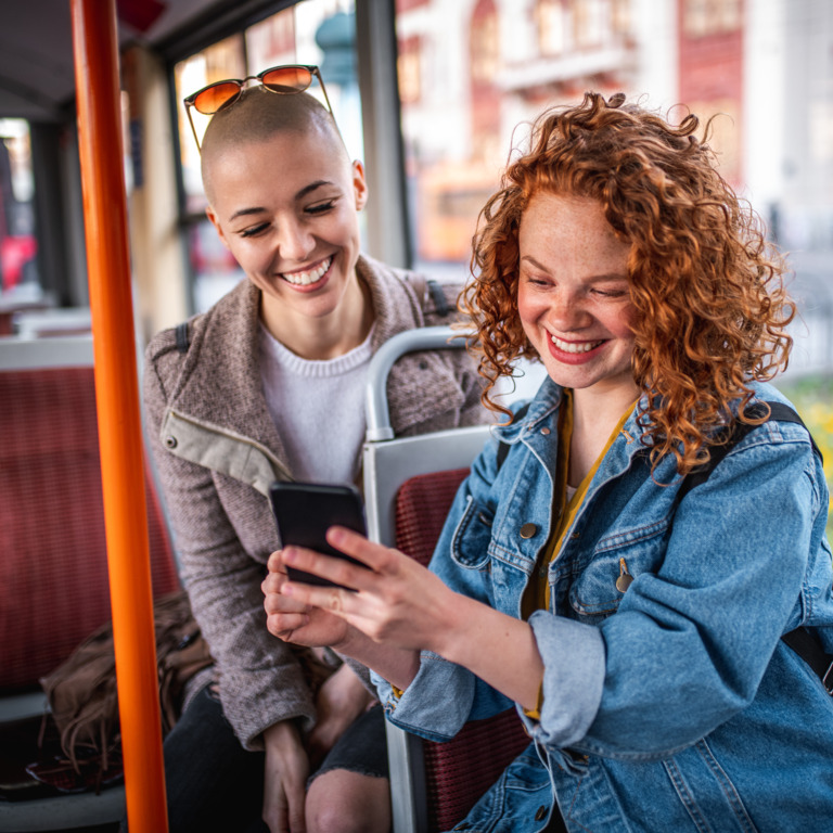 Zwei junge Frauen sitzen im Bus und schauen lachend auf ein Smartphone.