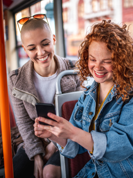 Zwei junge Frauen sitzen im Bus und schauen lachend auf ein Smartphone.
