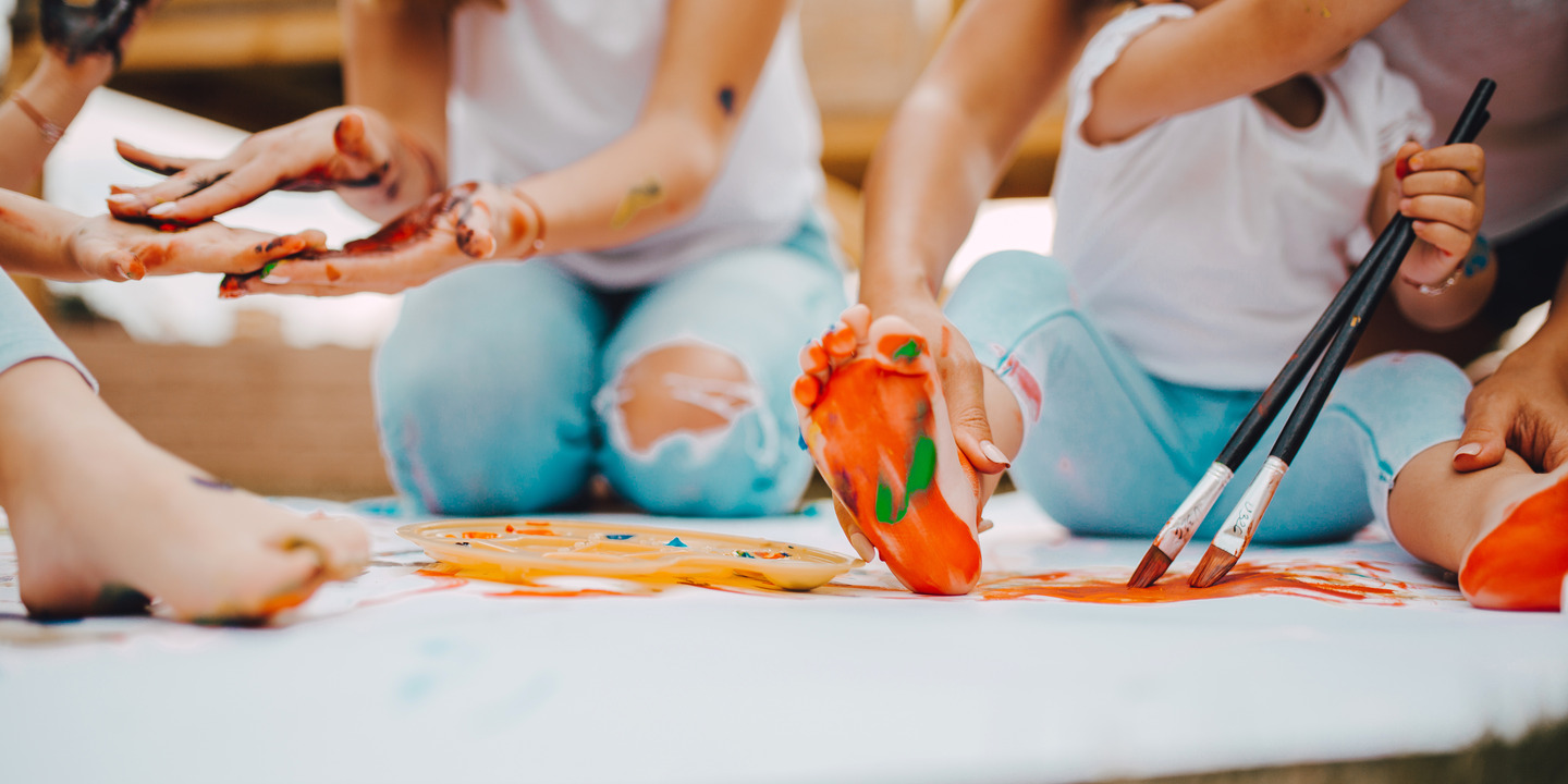 Kinder und Erzieher malen gemeinsam auf dem Fußboden. Die Fußsohle des Kindes ist orange mit grünen Tupfen angemalt.