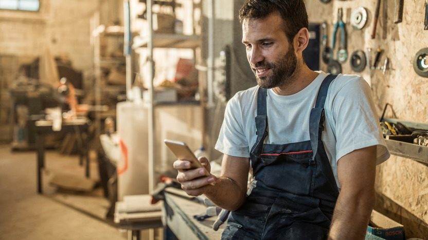Ein männlicher Handwerker in Arbeitskleidung sitzt auf einer Werkbank und schaut auf sein Smartphone
