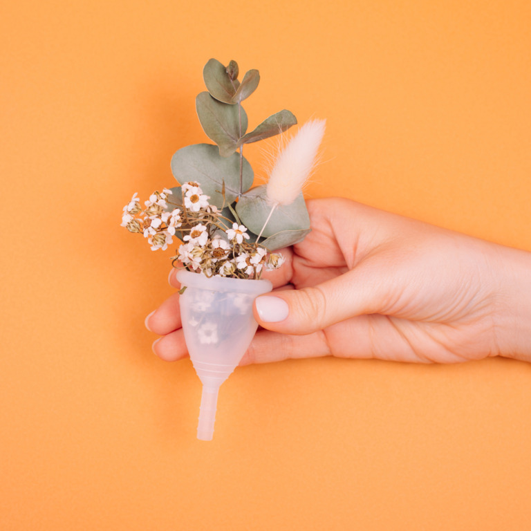 Hand hält eine Menstruationstasse mit Pflanzen vor einem orangenen Hintergrund.