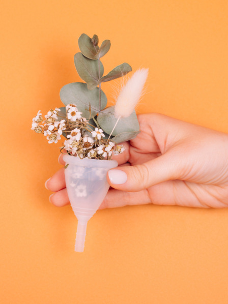 Weiße Menstruationstasse mit Blume in der Hand der Frau auf orangem Hintergrund