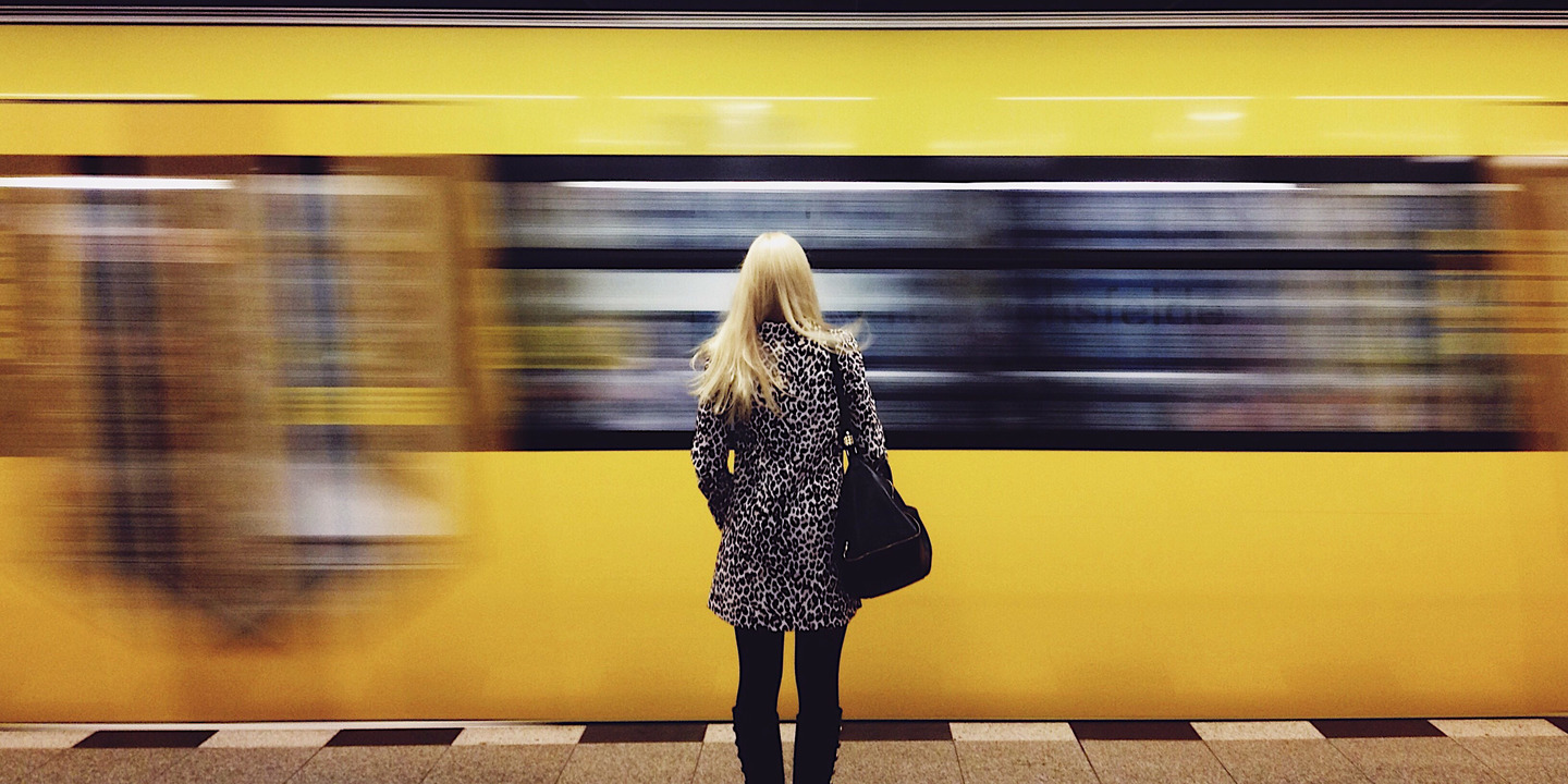 Blonde Frau steht rückwärts in U-Bahnhof vor gelber fahrender U-Bahn. 