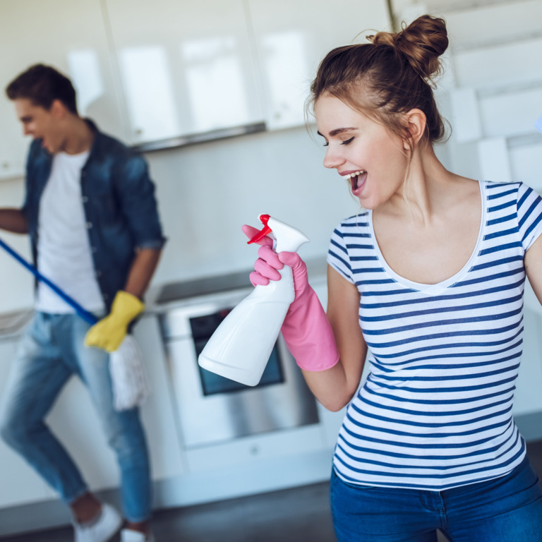 junger Mann mit Wischmopp und junge Frau mit Sprühflasche albern herum beim Kücheputzen