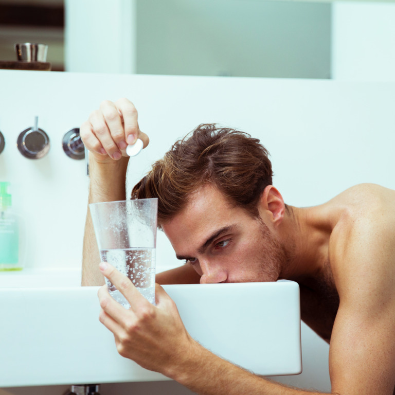 Junger Mann mit nackten Oberkörper stützt sich müde auf einem Waschbecken ab und wirft eine Kopfschmerztablette in ein Wasserglas. 