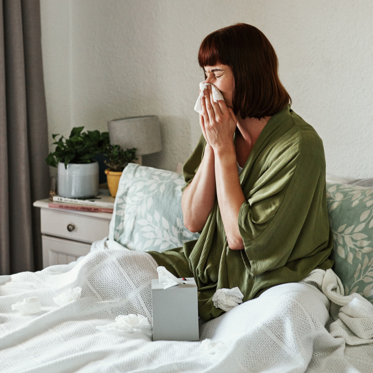 erkältete Frau sitzt im Bett und putzt ihre Nase