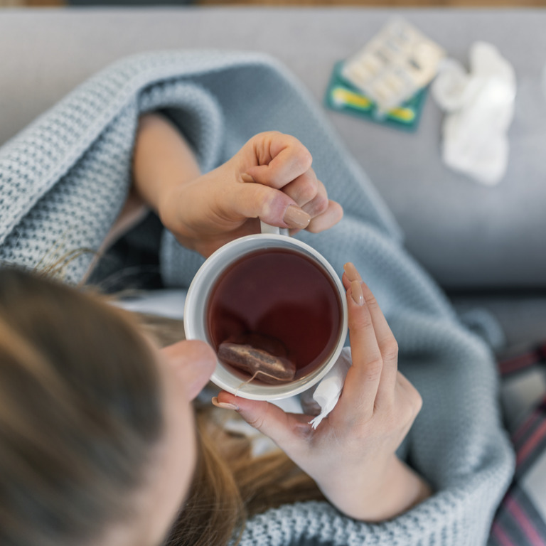 erkältete Frau sitzt eingehüllt in eine hellblaue Wolldecke auf dem Sofa und trinkt Tee