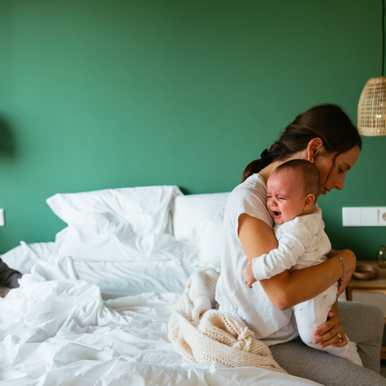 Unglücklich schauende Frau sitzt auf dem Bett und hält schreiendes Baby in den Armen.