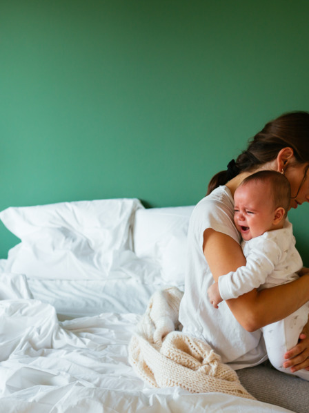 Mutter sitzt erschöpft auf dem Bett und tröstet ihr weinendes Baby, der zweijährige Bruder schaut zu