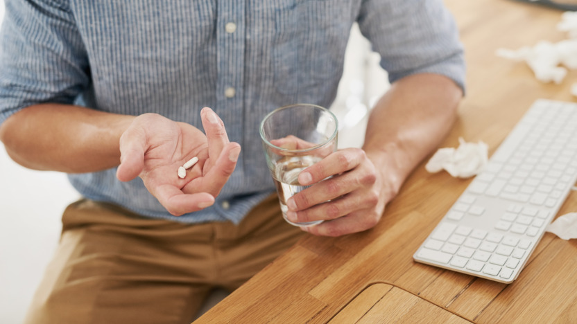 Mann sitzt am Schreibtisch und hält Tabletten und ein Glas Wasser in der Hand.