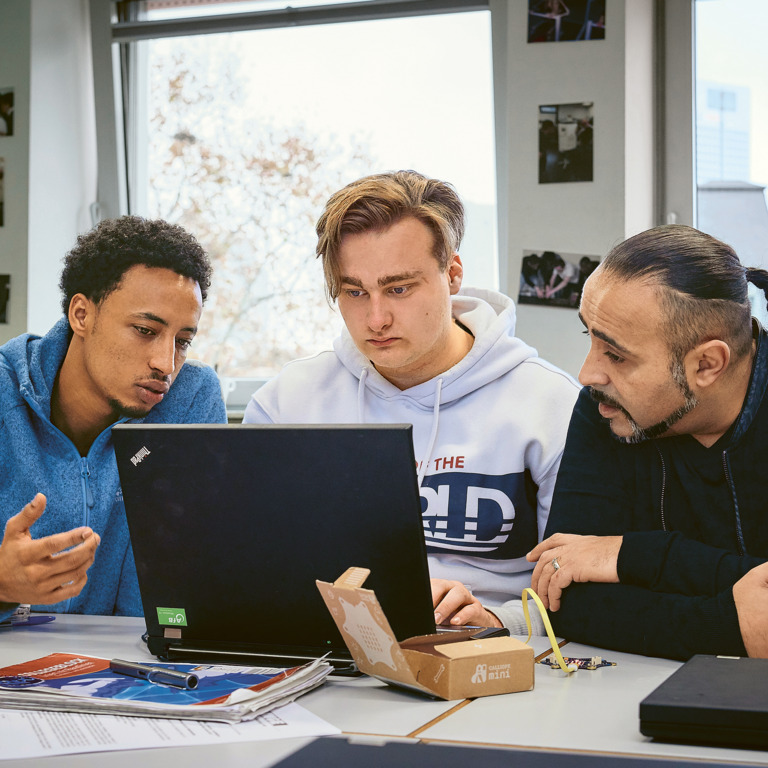 Kadim Tas von der Initiative "JOBLINGE" unterhält sich mit zwei Jugendlichen mit Migrationshintergrund vor einem Laptop.  