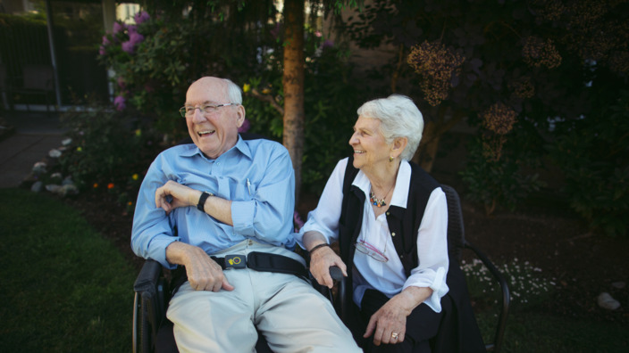 Seniorin und ihr pflegebedürftiger Mann sitzen gut gelaunt im Garten