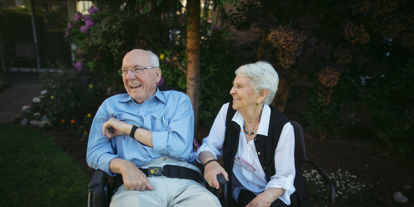 Seniorin und ihr pflegebedürftiger Mann sitzen gut gelaunt im Garten