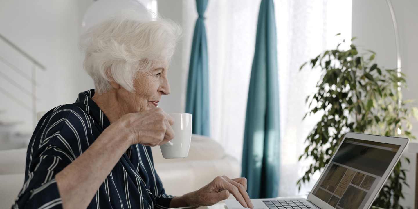 Seniorin sitzt mit einer Tasse Tee vor dem Laptop und prüft ihre Unterlagen
