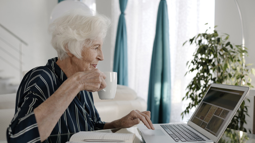 Ältere Frau sitzt vor dem Laptop mit einem Becher in der rechten Hand