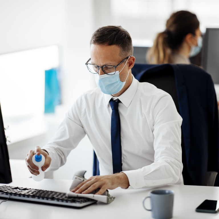 Angestellter sitzt mit Mundschutz vor seinem Büro-Rechner und desinfiziert ihn