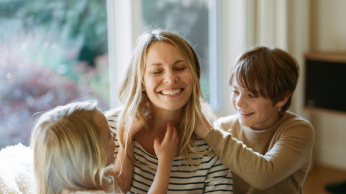 Junge Mutter sitzt lachend mit Kindern auf dem Sofa.