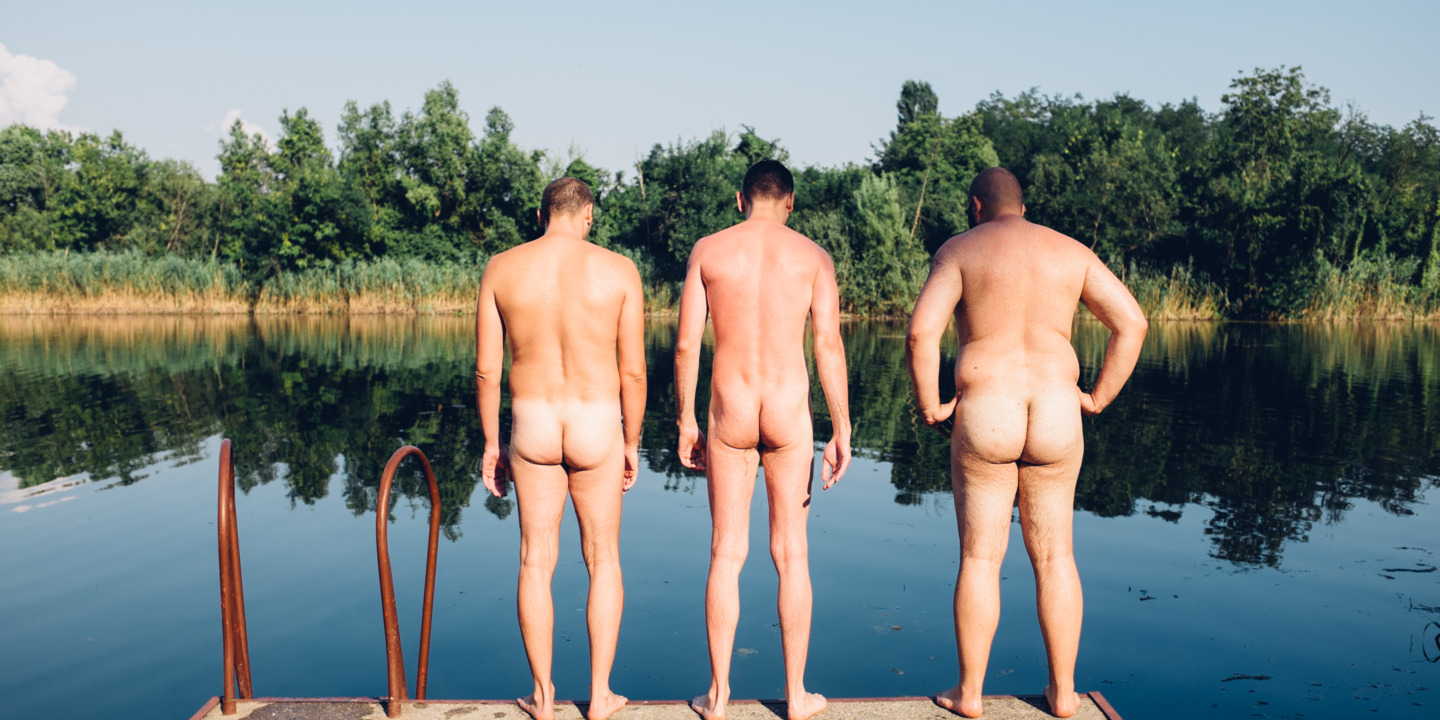 Drei nackte Männer stehen rückwärts auf Steg und blicken auf einen See. 