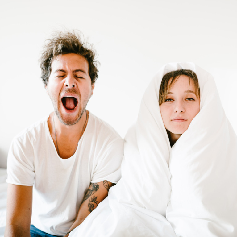 Gähnender Mann und Frau mit Bettdecke um den Kopf sitzen auf dem Bett