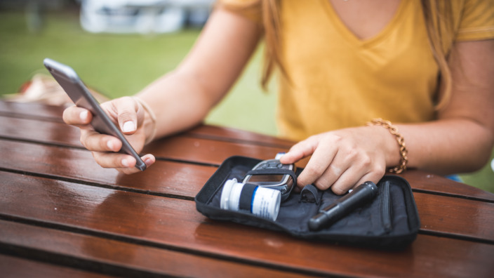 Frau sitzt mit geöffneter Diabetikertasche an einem Tisch und schaut auf ihr Handy